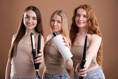 Photo of Beautiful women with different hair colors holding curling iron, shampoo and straightener on dark beige background