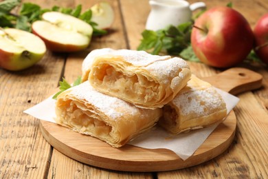 Photo of Pieces of tasty apple strudel with powdered sugar and fruits on wooden table, closeup