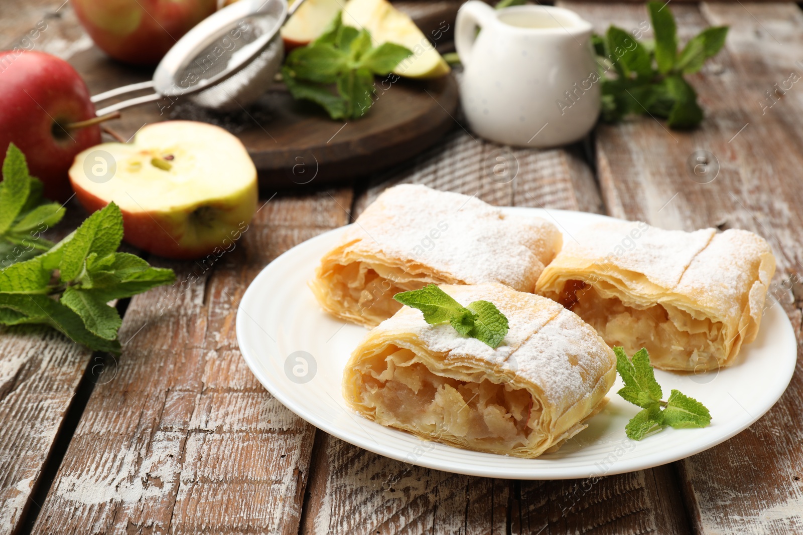 Pieces of tasty apple strudel with powdered sugar, mint and fruits on wooden table, closeup Photo of Pieces of tasty apple strudel with powdered sugar, mint and fruits on wooden table, closeup