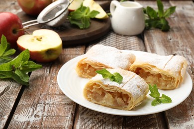 Photo of Pieces of tasty apple strudel with powdered sugar, mint and fruits on wooden table, closeup