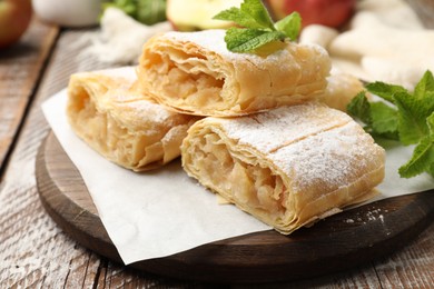 Photo of Pieces of tasty apple strudel with powdered sugar, mint and fruits on wooden table, closeup