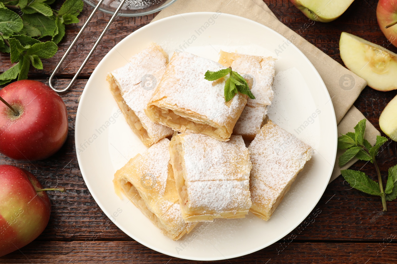 Pieces of tasty apple strudel with powdered sugar, mint and fruits on wooden table, flat lay Photo of Pieces of tasty apple strudel with powdered sugar, mint and fruits on wooden table, flat lay