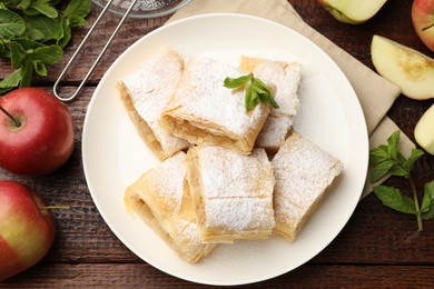 Photo of Pieces of tasty apple strudel with powdered sugar, mint and fruits on wooden table, flat lay