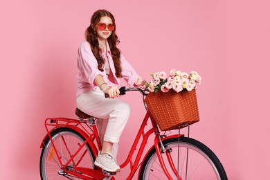Photo of Beautiful teenage girl in sunglasses riding bicycle with basket of flowers on pink background