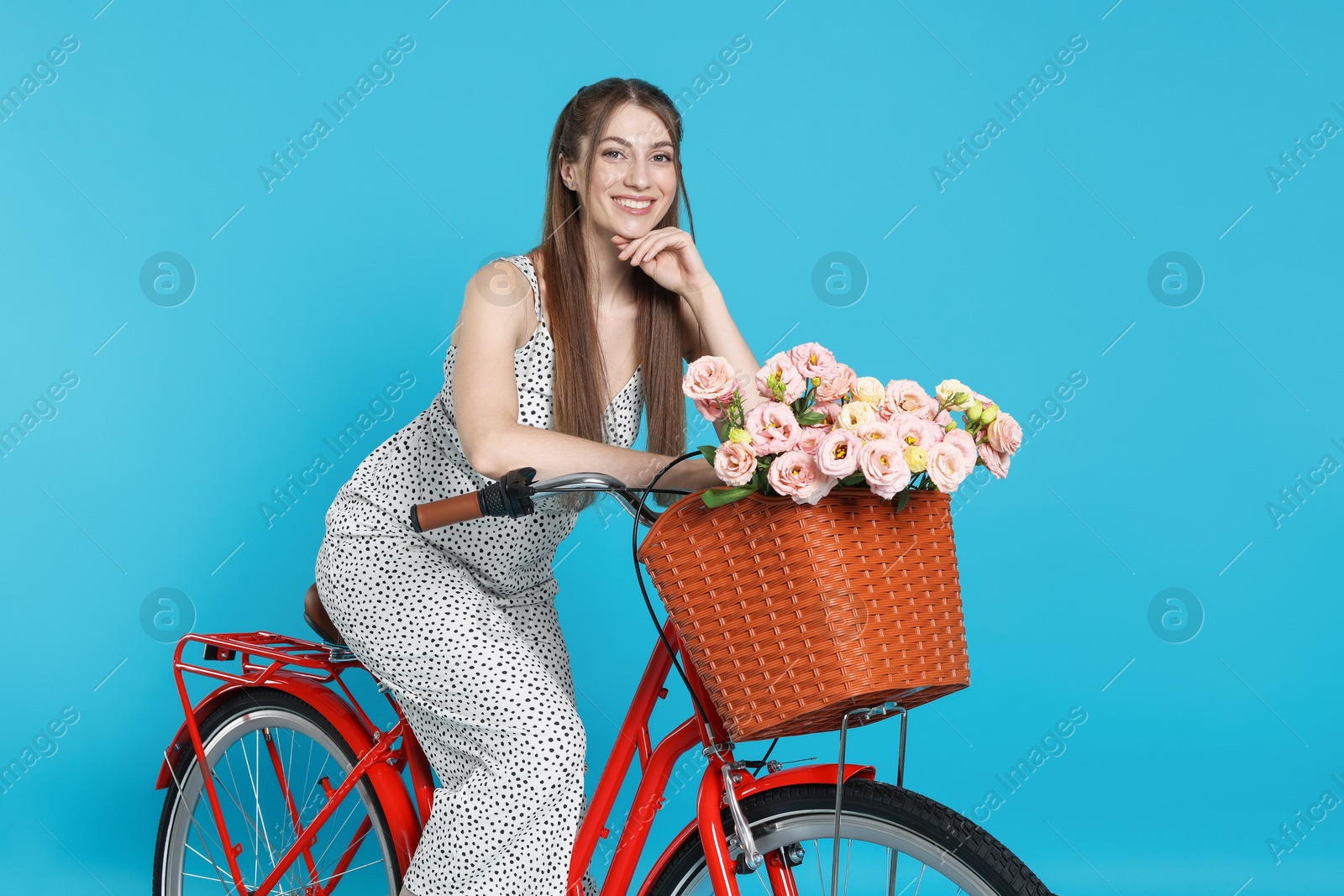 Photo of Smiling woman posing on bicycle with basket of flowers against light blue background