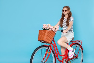 Photo of Beautiful teenage girl in sunglasses riding bicycle with basket of flowers against light blue background. Space for text