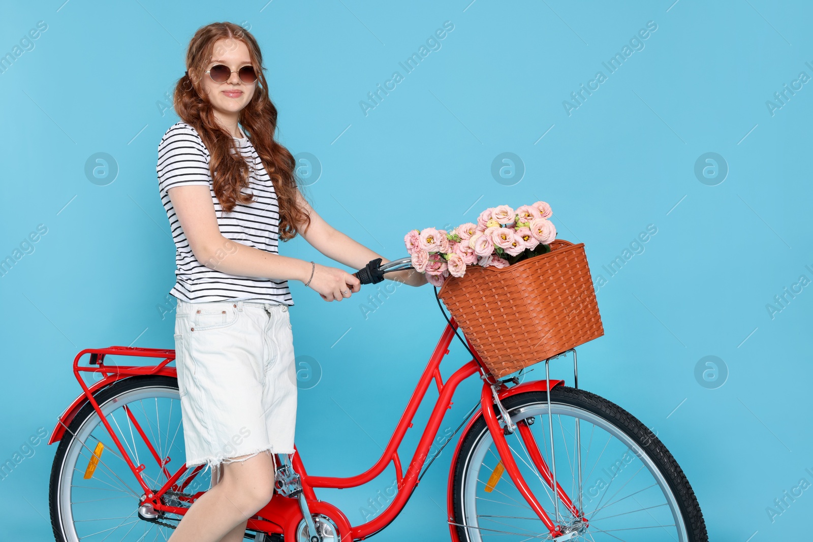 Beautiful teenage girl in sunglasses and bicycle with basket of flowers against light blue background Photo of Beautiful teenage girl in sunglasses and bicycle with basket of flowers against light blue background