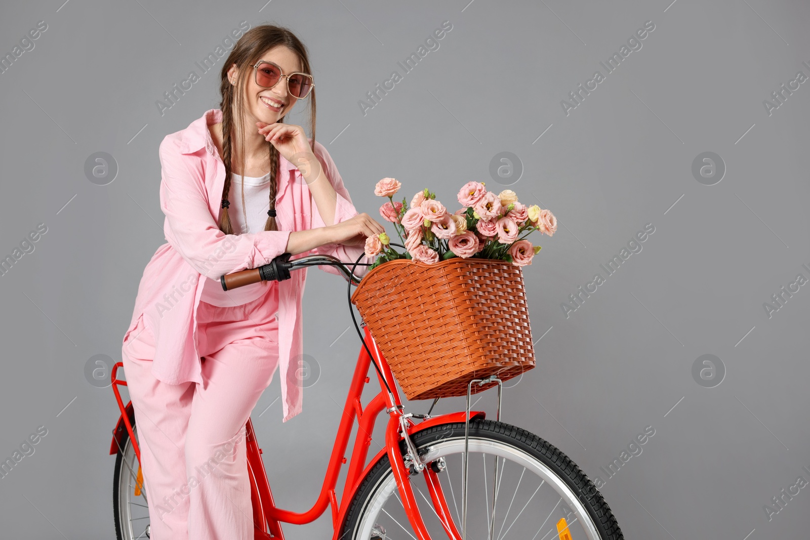 Smiling woman in sunglasses and bicycle with basket of flowers against grey background Photo of Smiling woman in sunglasses and bicycle with basket of flowers against grey background