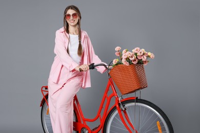 Photo of Smiling woman in sunglasses and bicycle with basket of flowers against grey background