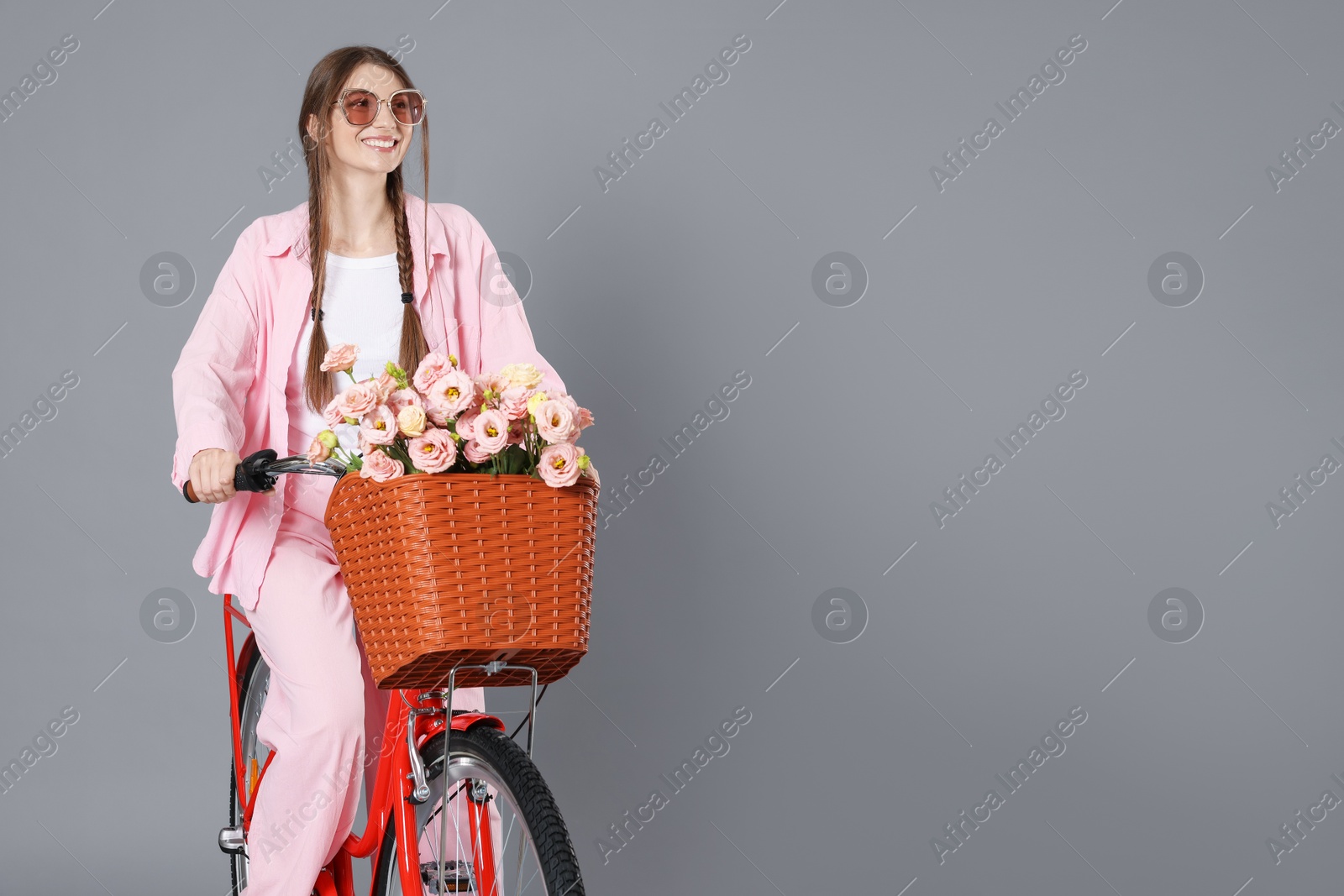 Smiling woman in sunglasses riding bicycle with basket of flowers against grey background. Space for text Photo of Smiling woman in sunglasses riding bicycle with basket of flowers against grey background. Space for text