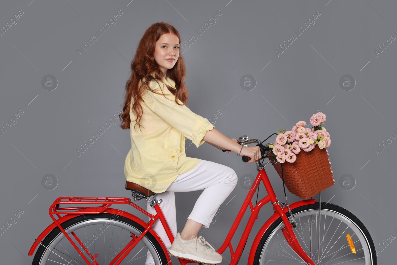 Beautiful teenage girl riding bicycle with basket of flowers against grey background Photo of Beautiful teenage girl riding bicycle with basket of flowers against grey background