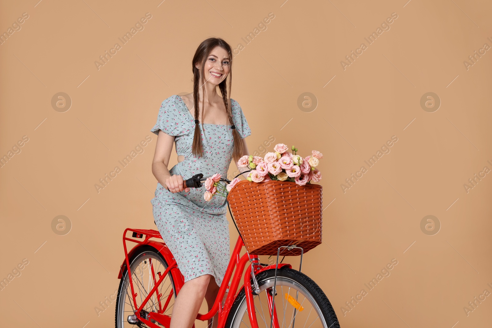 Smiling woman in dress riding bicycle with basket of flowers on beige background Photo of Smiling woman in dress riding bicycle with basket of flowers on beige background