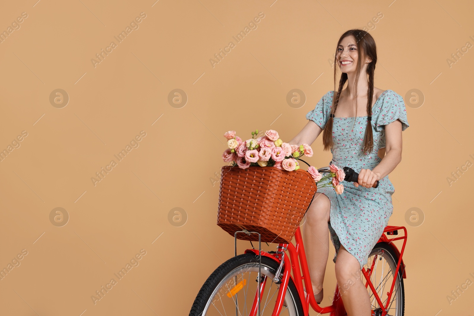 Smiling woman in dress riding bicycle with basket of flowers on beige background. Space for text Photo of Smiling woman in dress riding bicycle with basket of flowers on beige background. Space for text