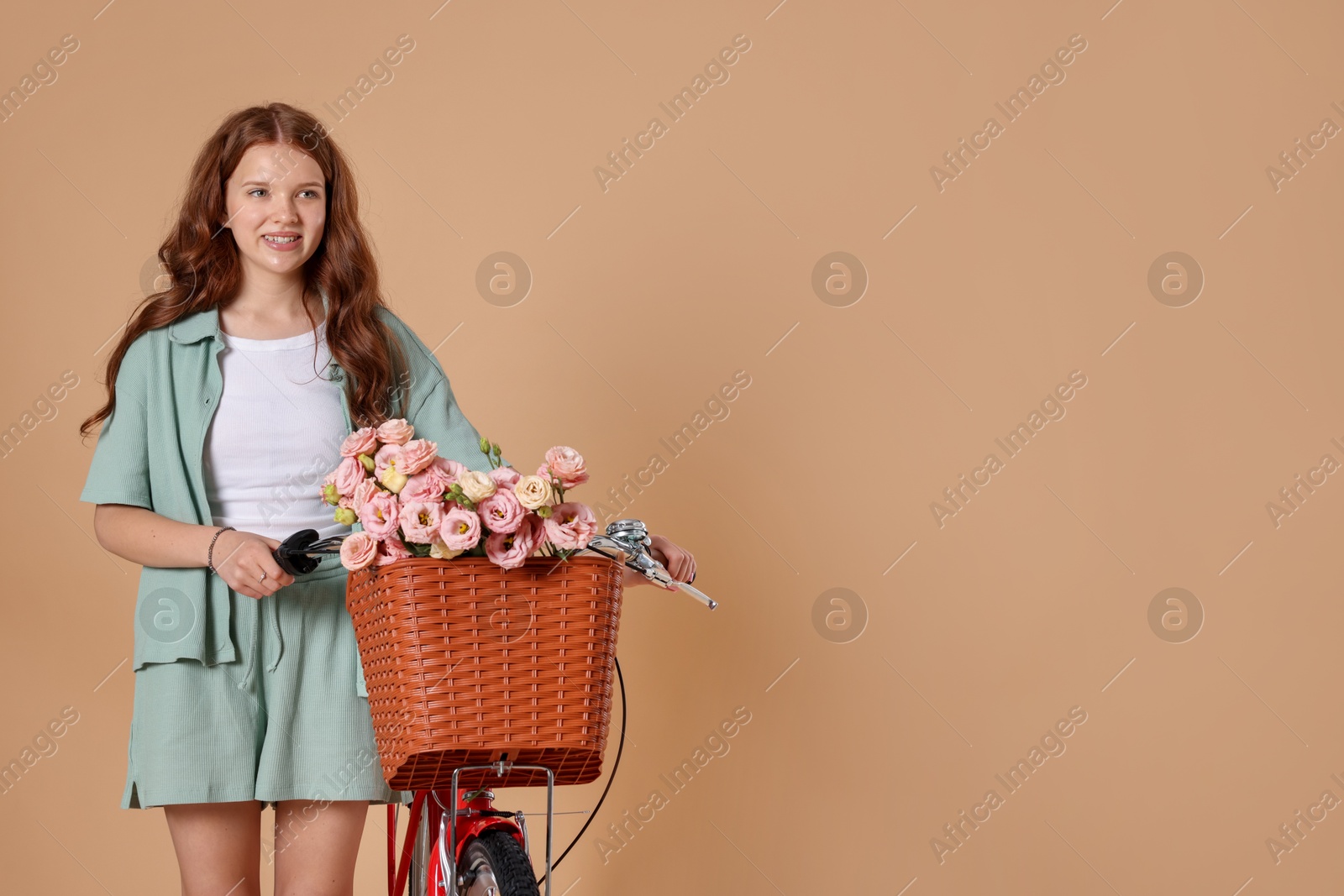 Smiling teenage girl and bicycle with basket of flowers against beige background. Space for text Photo of Smiling teenage girl and bicycle with basket of flowers against beige background. Space for text