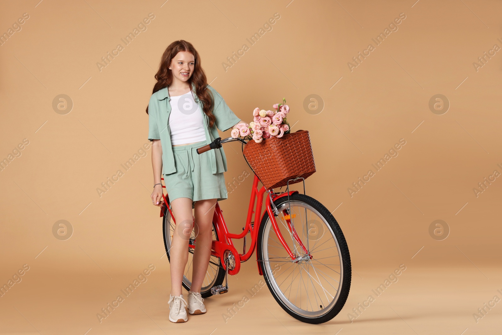 Smiling teenage girl and bicycle with basket of flowers against beige background Photo of Smiling teenage girl and bicycle with basket of flowers against beige background