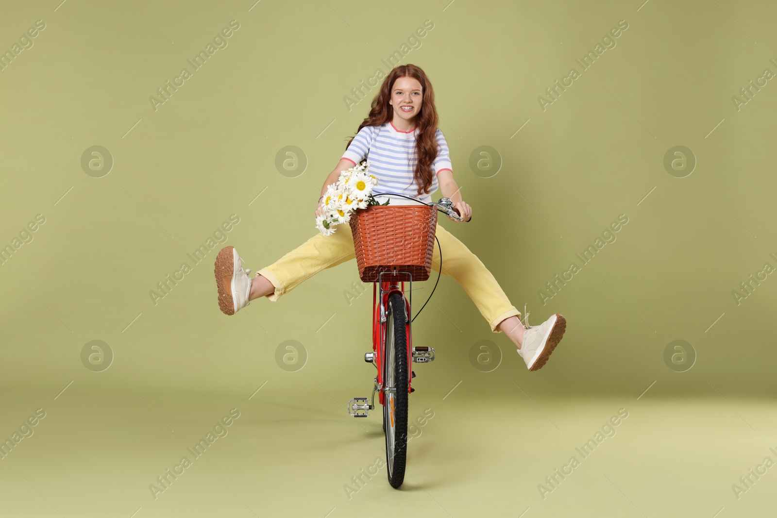 Smiling teenage girl riding bicycle with basket of flowers against green background Photo of Smiling teenage girl riding bicycle with basket of flowers against green background