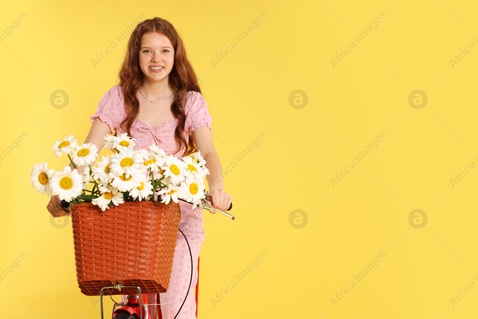 Smiling teenage girl riding bicycle with basket of flowers against yellow background. Space for text Photo of Smiling teenage girl riding bicycle with basket of flowers against yellow background. Space for text