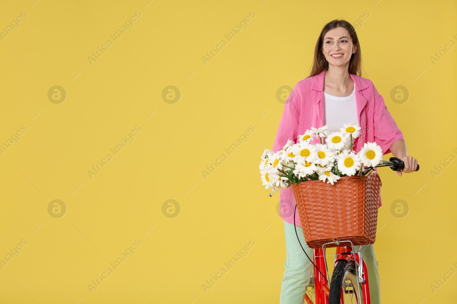 Smiling woman riding bicycle with basket of flowers against yellow background. Space for text Photo of Smiling woman riding bicycle with basket of flowers against yellow background. Space for text
