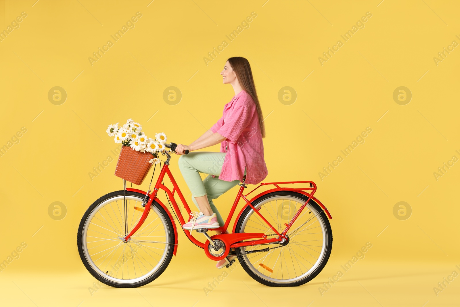 Smiling woman riding bicycle with basket of flowers against yellow background Photo of Smiling woman riding bicycle with basket of flowers against yellow background
