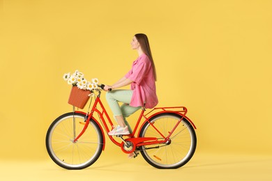 Photo of Smiling woman riding bicycle with basket of flowers against yellow background