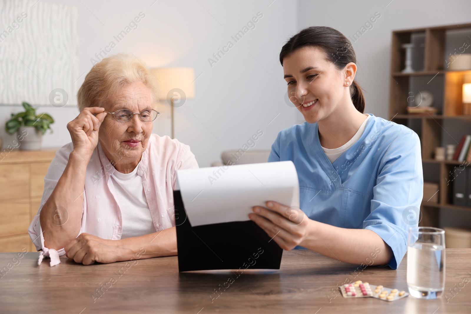 Photo of Nurse with clipboard examining senior woman at wooden table indoors. Home health care service