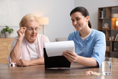 Photo of Nurse with clipboard examining senior woman at wooden table indoors. Home health care service