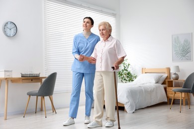 Photo of Nurse assisting senior woman with walking cane indoors. Home health care service