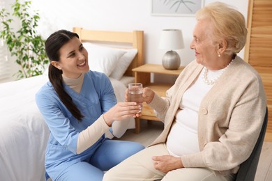 Photo of Nurse giving water to senior woman on sofa indoors. Home health care service