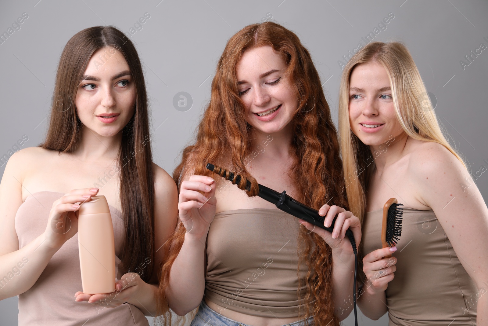 Beautiful women with different hair colors holding curling iron, shampoo and brush on grey background Photo of Beautiful women with different hair colors holding curling iron, shampoo and brush on grey background