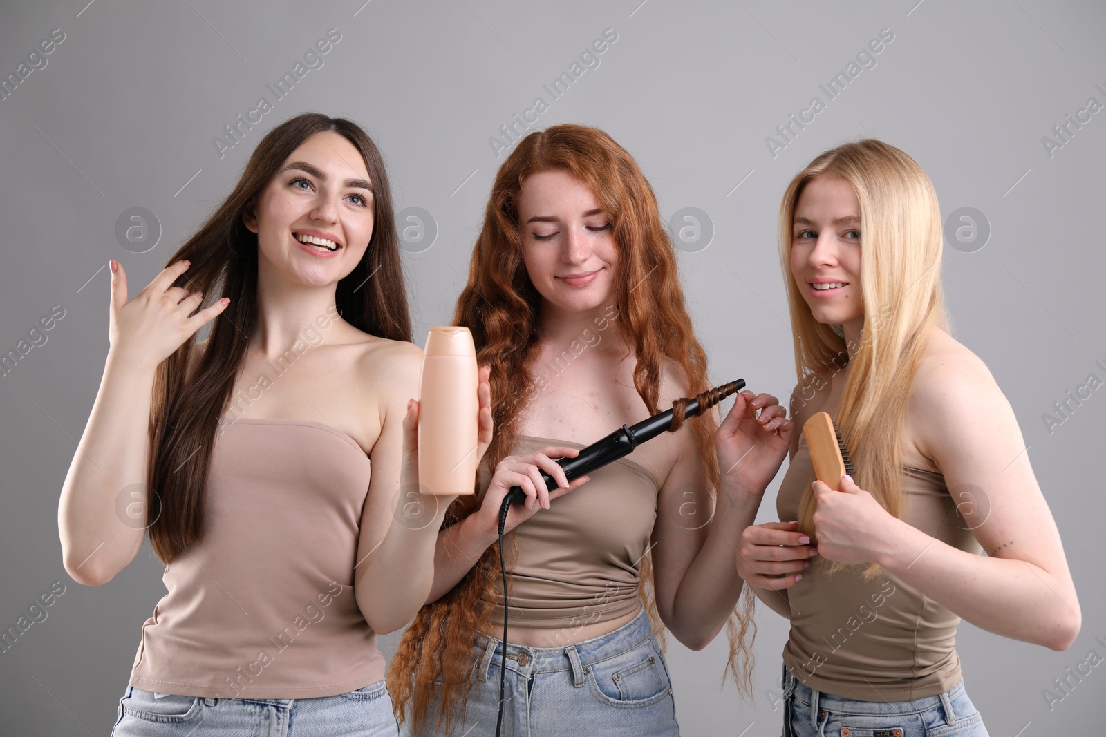Beautiful women with different hair colors holding curling iron, shampoo and brush on grey background Photo of Beautiful women with different hair colors holding curling iron, shampoo and brush on grey background