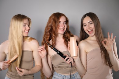 Photo of Beautiful women with different hair colors holding curling iron, shampoo and brush on grey background
