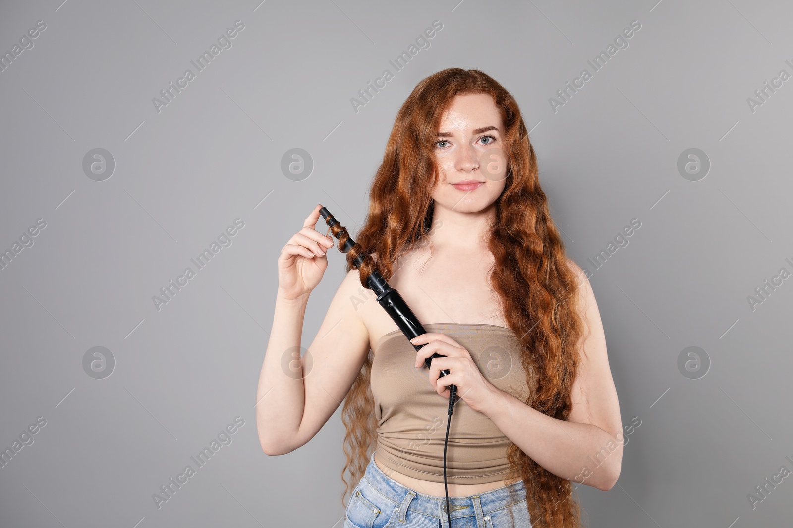 Beautiful woman with long wavy hair and curling iron on grey background, space for text Photo of Beautiful woman with long wavy hair and curling iron on grey background, space for text
