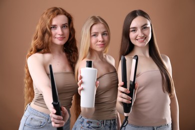 Photo of Beautiful women with different hair colors holding curling iron, shampoo and straightener on dark beige background