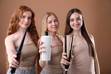 Photo of Beautiful women with different hair colors holding curling iron, shampoo and straightener on dark beige background
