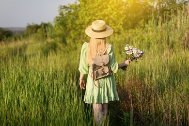 Photo of Woman with beautiful wildflowers, hat and backpack in meadow on sunny day, back view