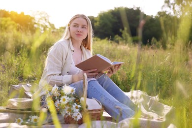 Photo of Woman with bouquet of wildflowers reading book on blanket in meadow