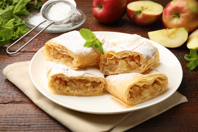 Photo of Pieces of tasty apple strudel with powdered sugar, mint and fruits on wooden table, closeup