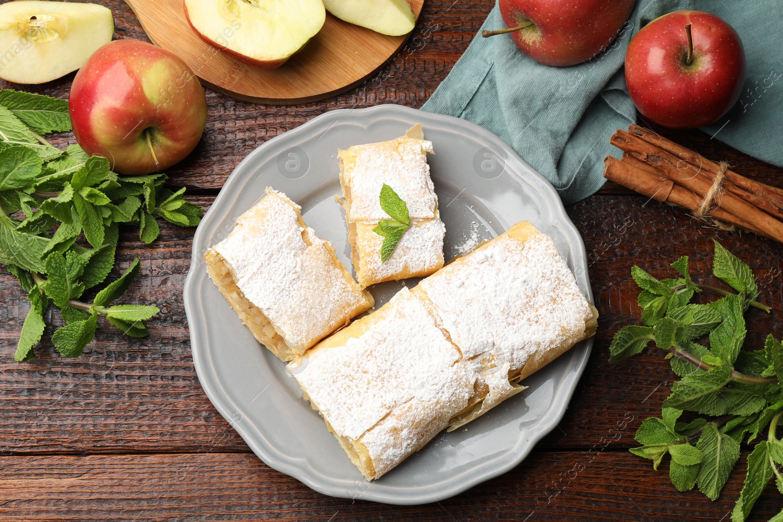 Pieces of tasty apple strudel with powdered sugar, mint and fruits on wooden table, flat lay Photo of Pieces of tasty apple strudel with powdered sugar, mint and fruits on wooden table, flat lay