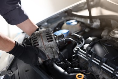 Photo of Auto part. Professional mechanic with alternator near open hood of car indoors, closeup