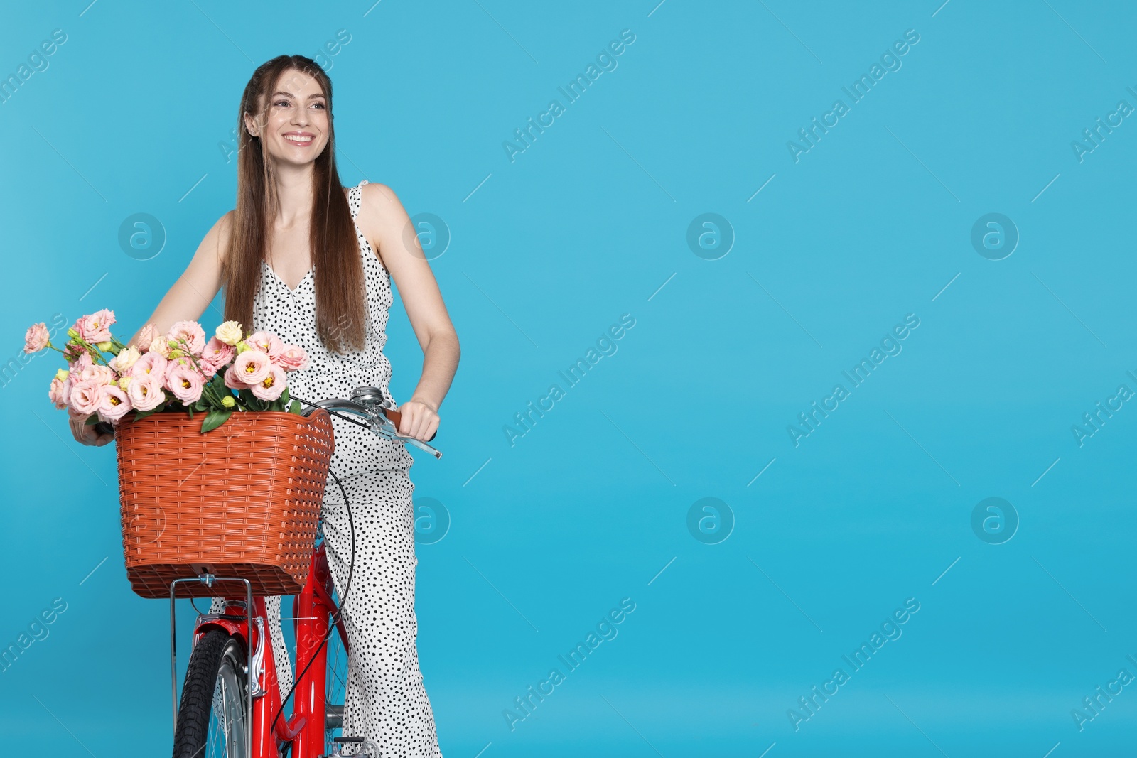 Photo of Smiling woman riding bicycle with basket of flowers against light blue background. Space for text