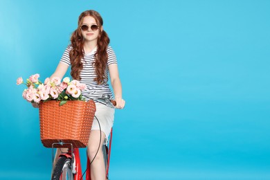 Photo of Beautiful teenage girl in sunglasses riding bicycle with basket of flowers against light blue background. Space for text