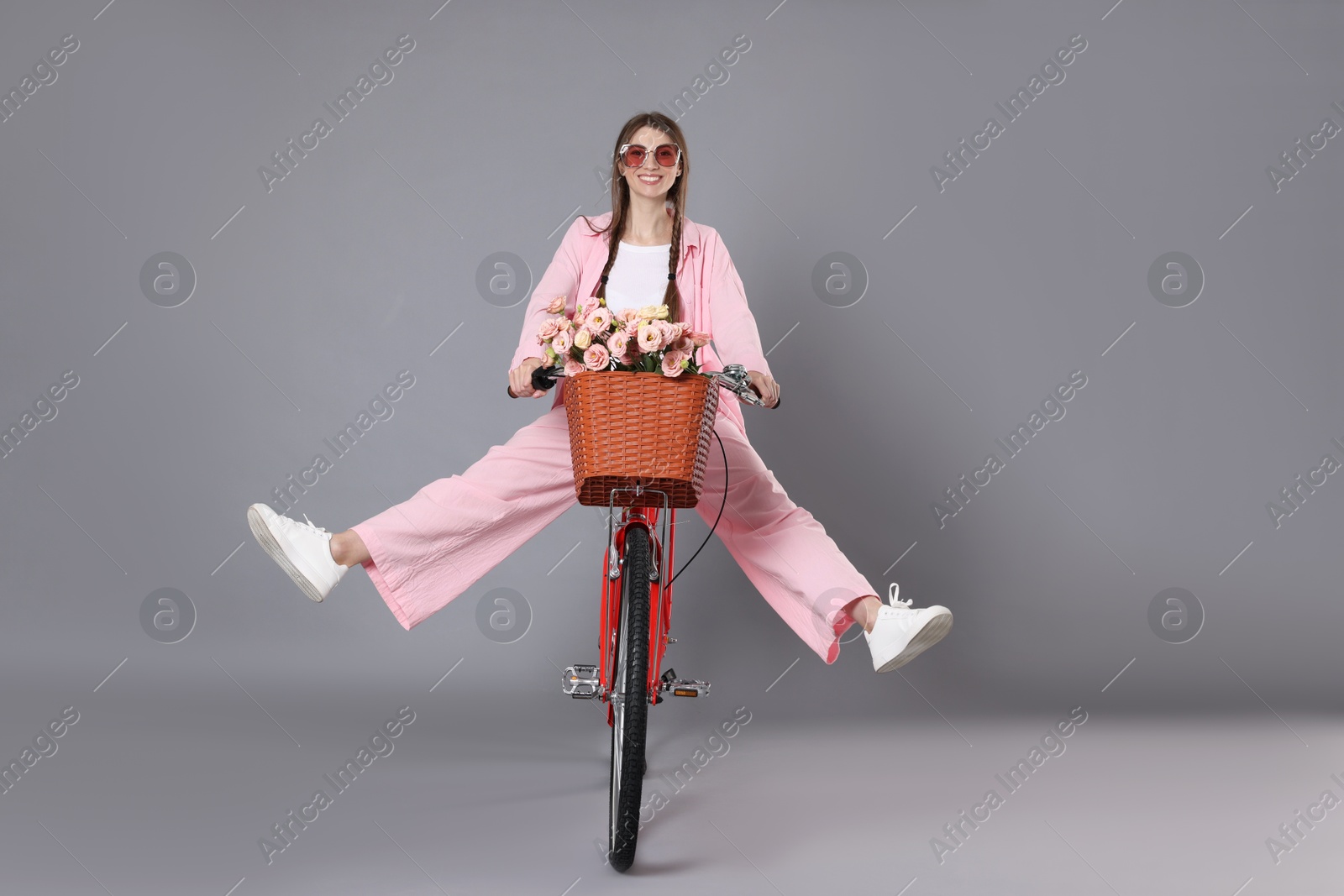 Smiling woman in sunglasses riding bicycle with basket of flowers against grey background Photo of Smiling woman in sunglasses riding bicycle with basket of flowers against grey background