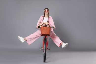 Photo of Smiling woman in sunglasses riding bicycle with basket of flowers against grey background