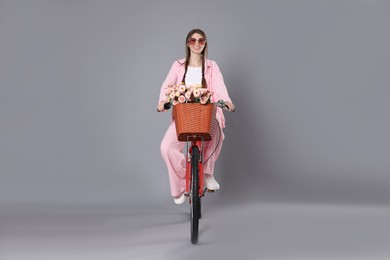 Photo of Smiling woman in sunglasses riding bicycle with basket of flowers against grey background