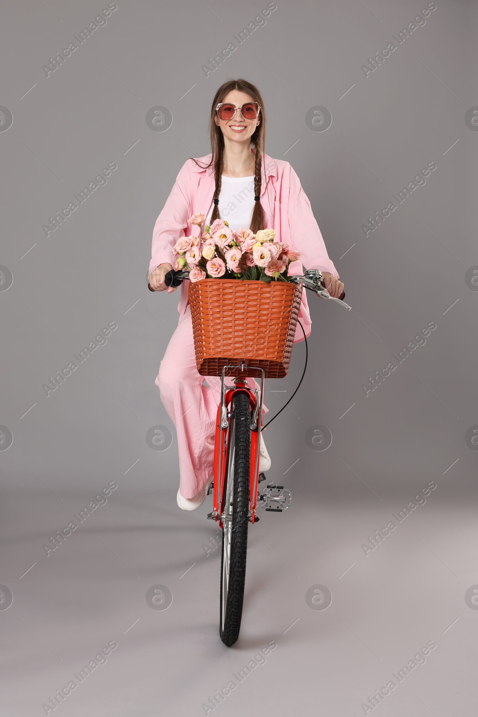 Photo of Smiling woman in sunglasses riding bicycle with basket of flowers against grey background