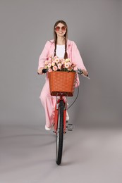 Photo of Smiling woman in sunglasses riding bicycle with basket of flowers against grey background
