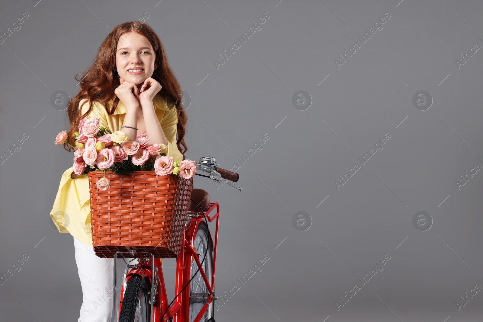 Smiling teenage girl and bicycle with basket of flowers against grey background. Space for text Photo of Smiling teenage girl and bicycle with basket of flowers against grey background. Space for text