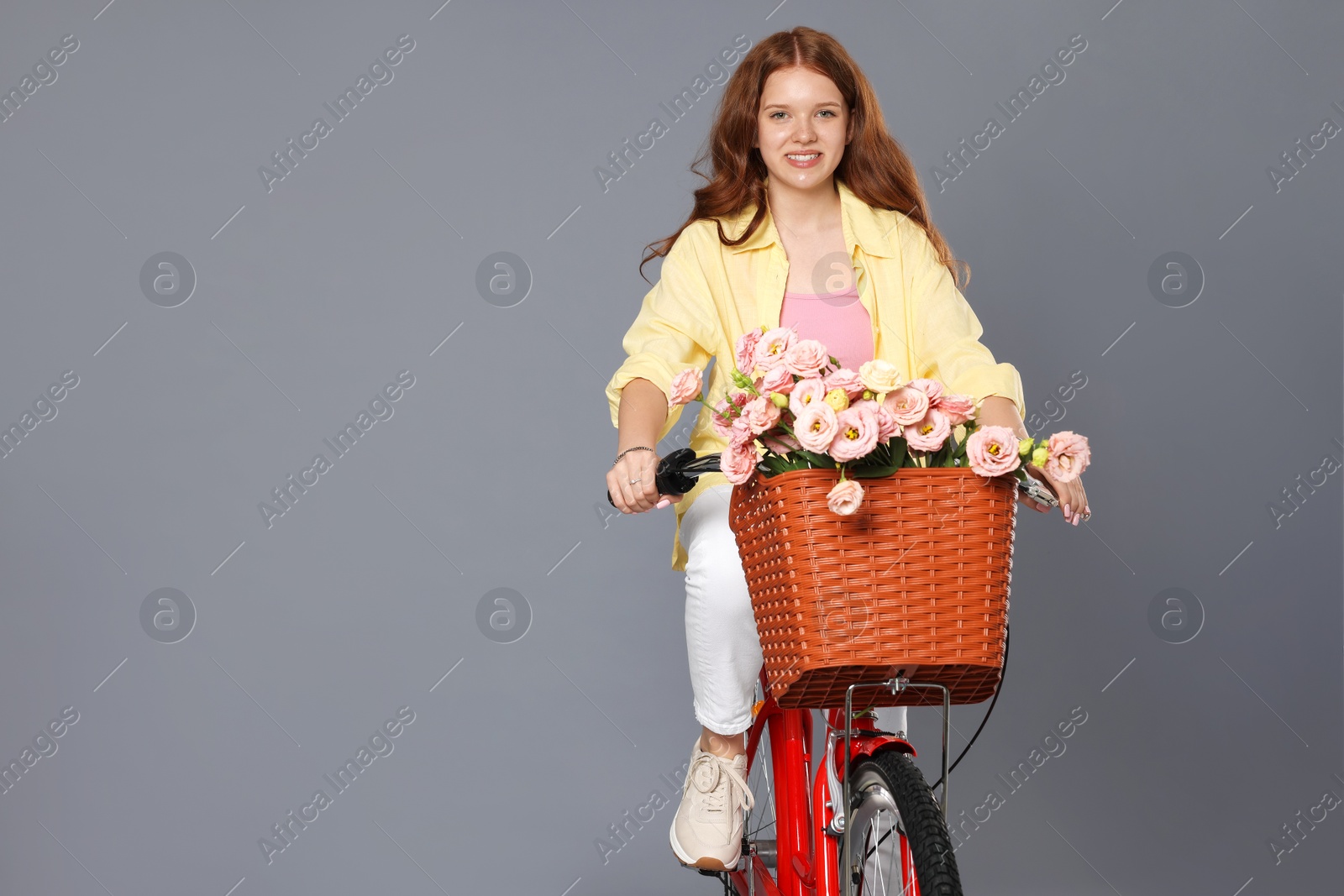 Smiling teenage girl riding bicycle with basket of flowers against grey background. Space for text Photo of Smiling teenage girl riding bicycle with basket of flowers against grey background. Space for text