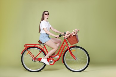 Photo of Smiling woman in sunglasses riding bicycle with basket of flowers on green background