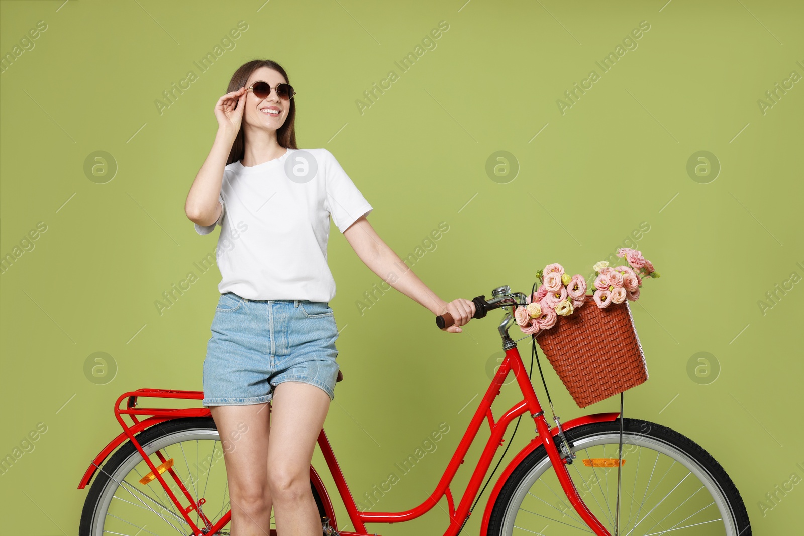 Smiling woman in sunglasses and bicycle with basket of flowers on green background Photo of Smiling woman in sunglasses and bicycle with basket of flowers on green background